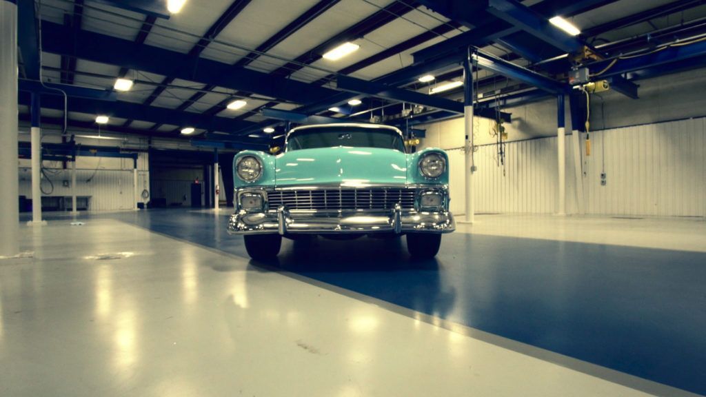 Teal vintage car parked in a bright, empty garage with blue and gray floors.