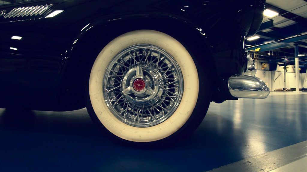 Close-up of a black vintage car with whitewall tire, chrome wire wheel, and red center cap on blue floor.