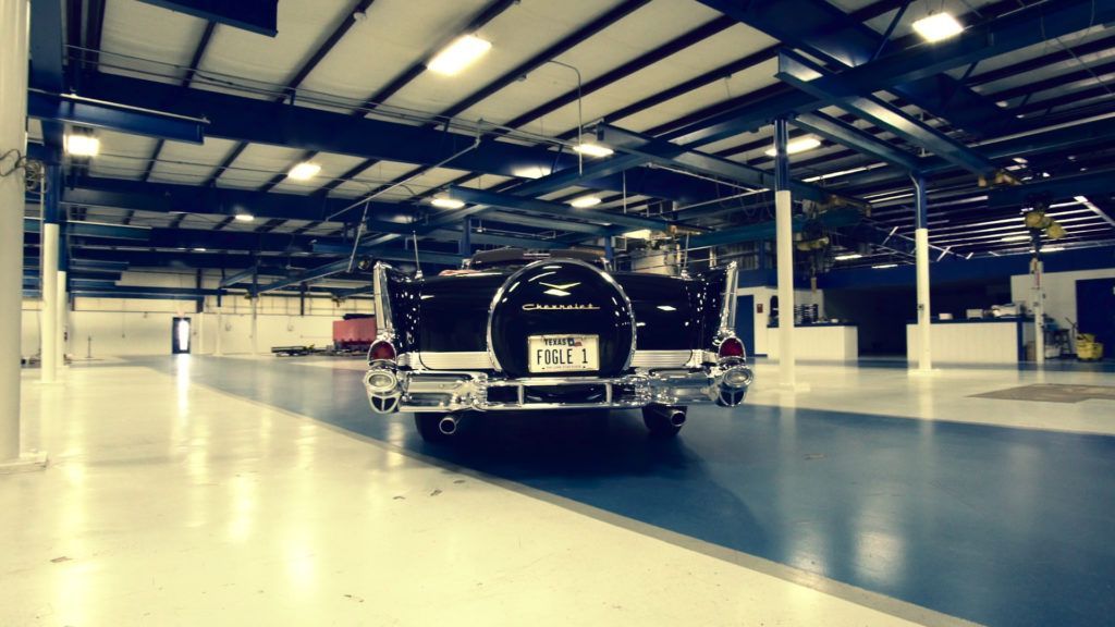 Black classic car driving inside a large industrial building with bright lighting.