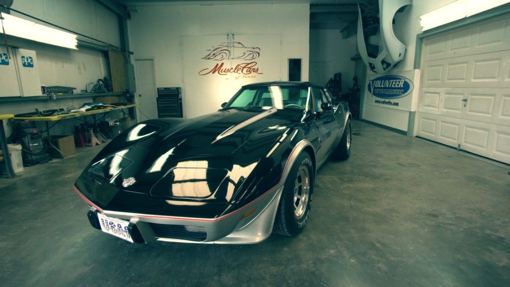 Black Corvette in a garage; bright lighting reflects off the car's surface.