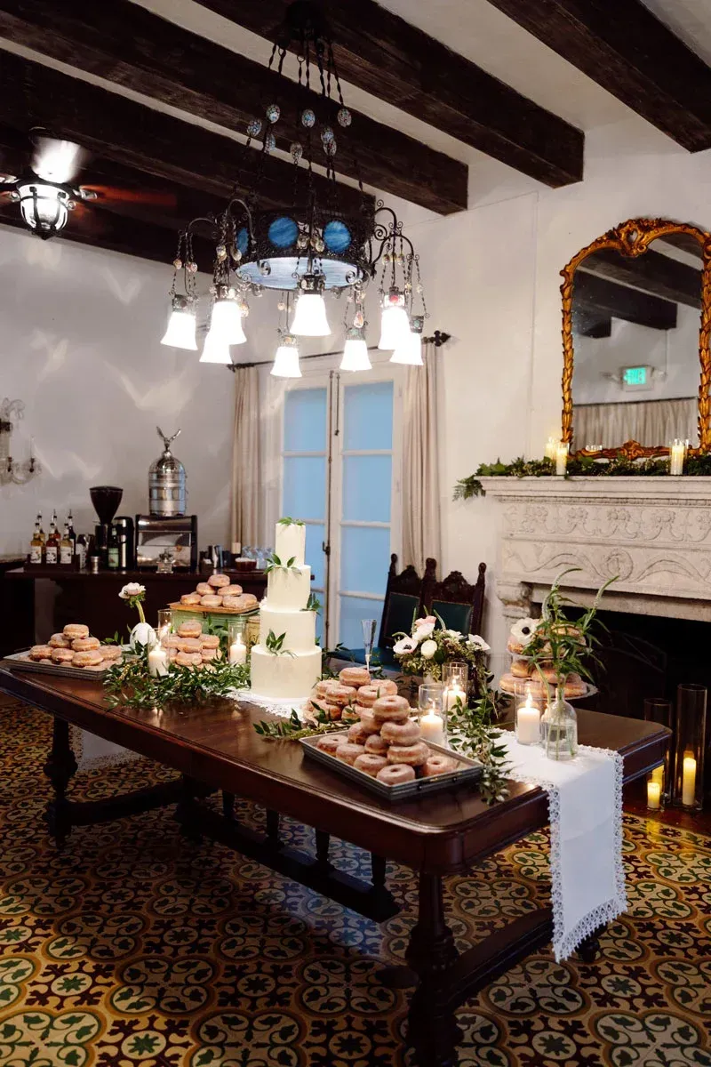 Wedding cake and desserts on a long wooden table in a room with a fireplace and chandelier.