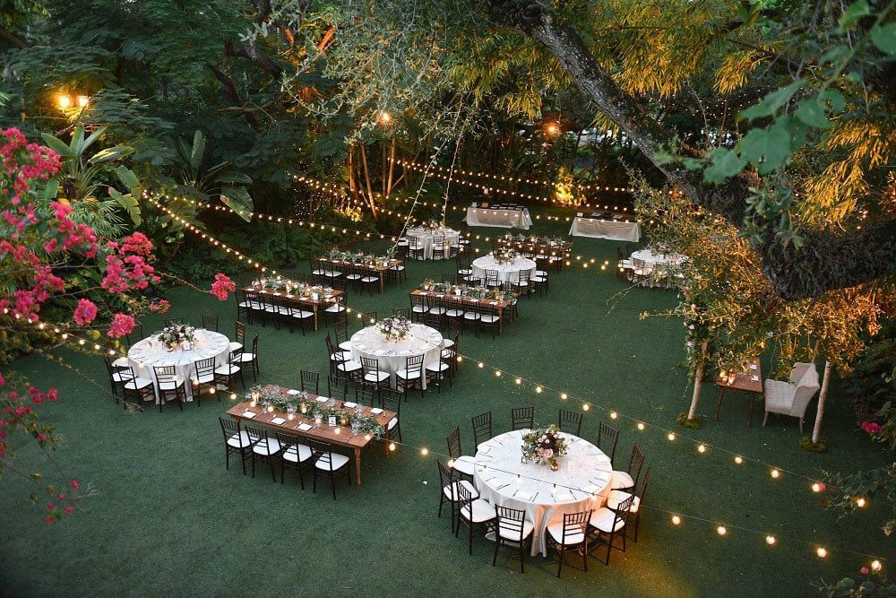 Outdoor reception with tables set for guests, strung with lights, in a garden.