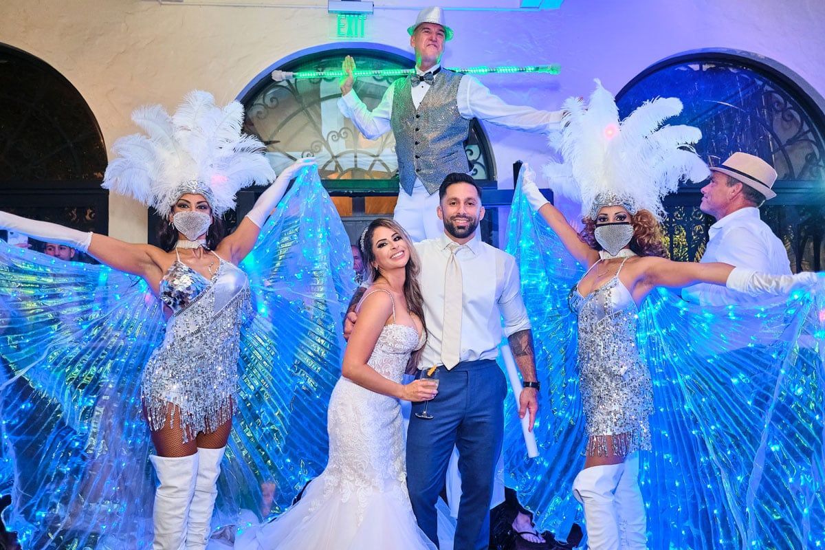 Bride and groom pose with costumed performers at an outdoor event with blue lighting.
