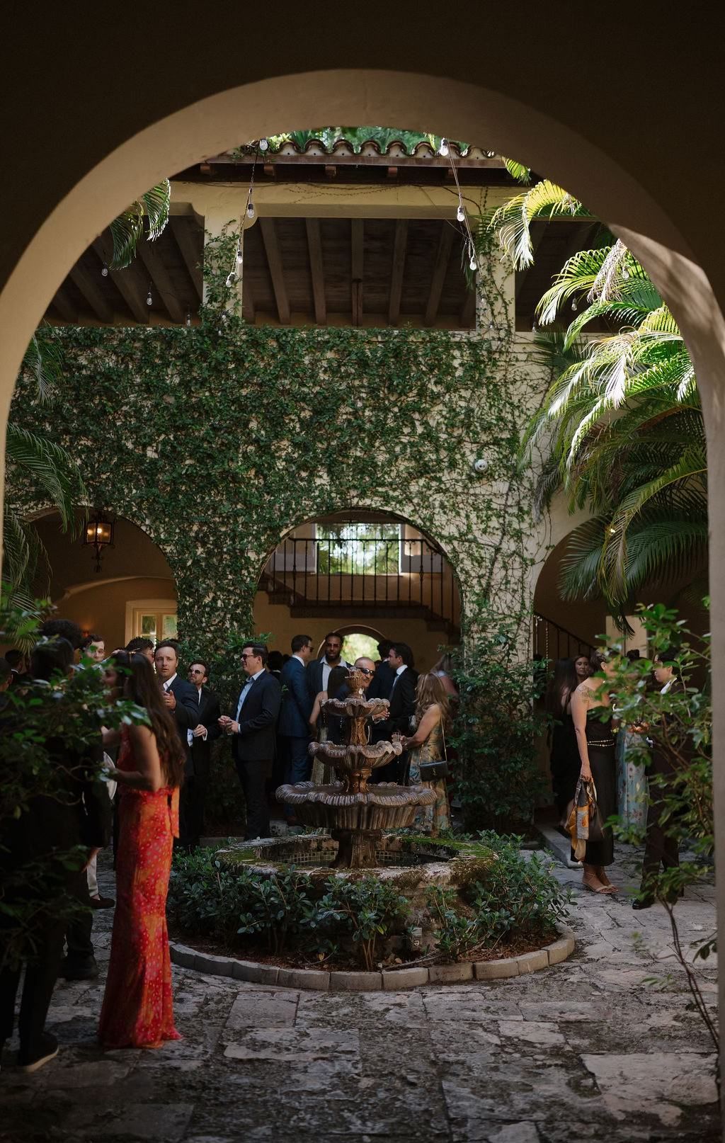 Courtyard gathering, stone fountain, ivy-covered walls, people in formal attire.