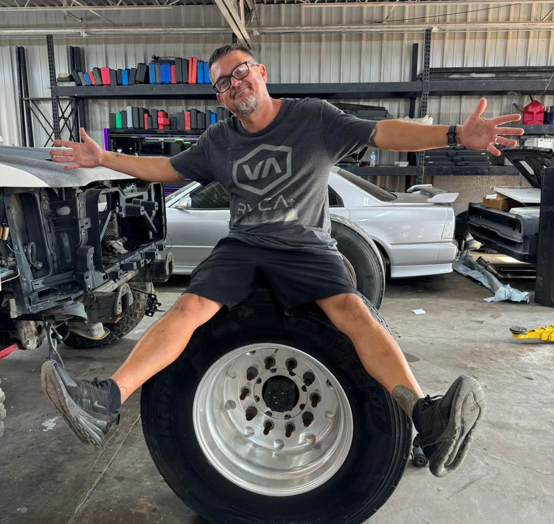 Man sitting on a large tire in a workshop, arms outstretched with a smile.