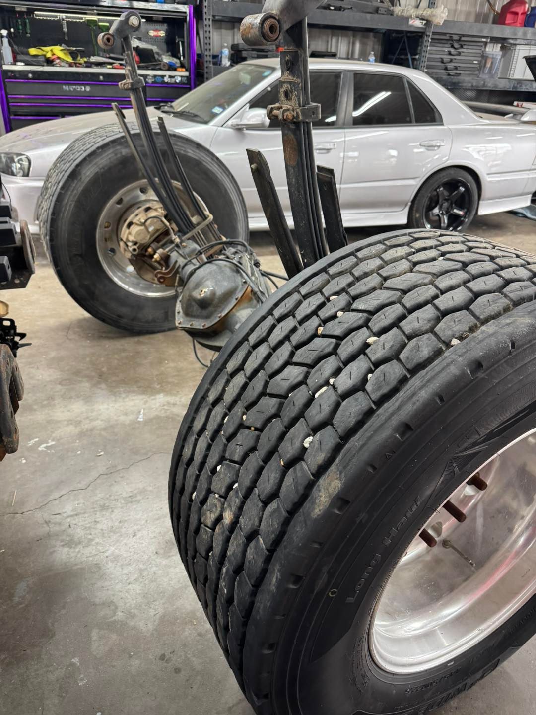 Truck axle with large tires in a garage, silver car in background.