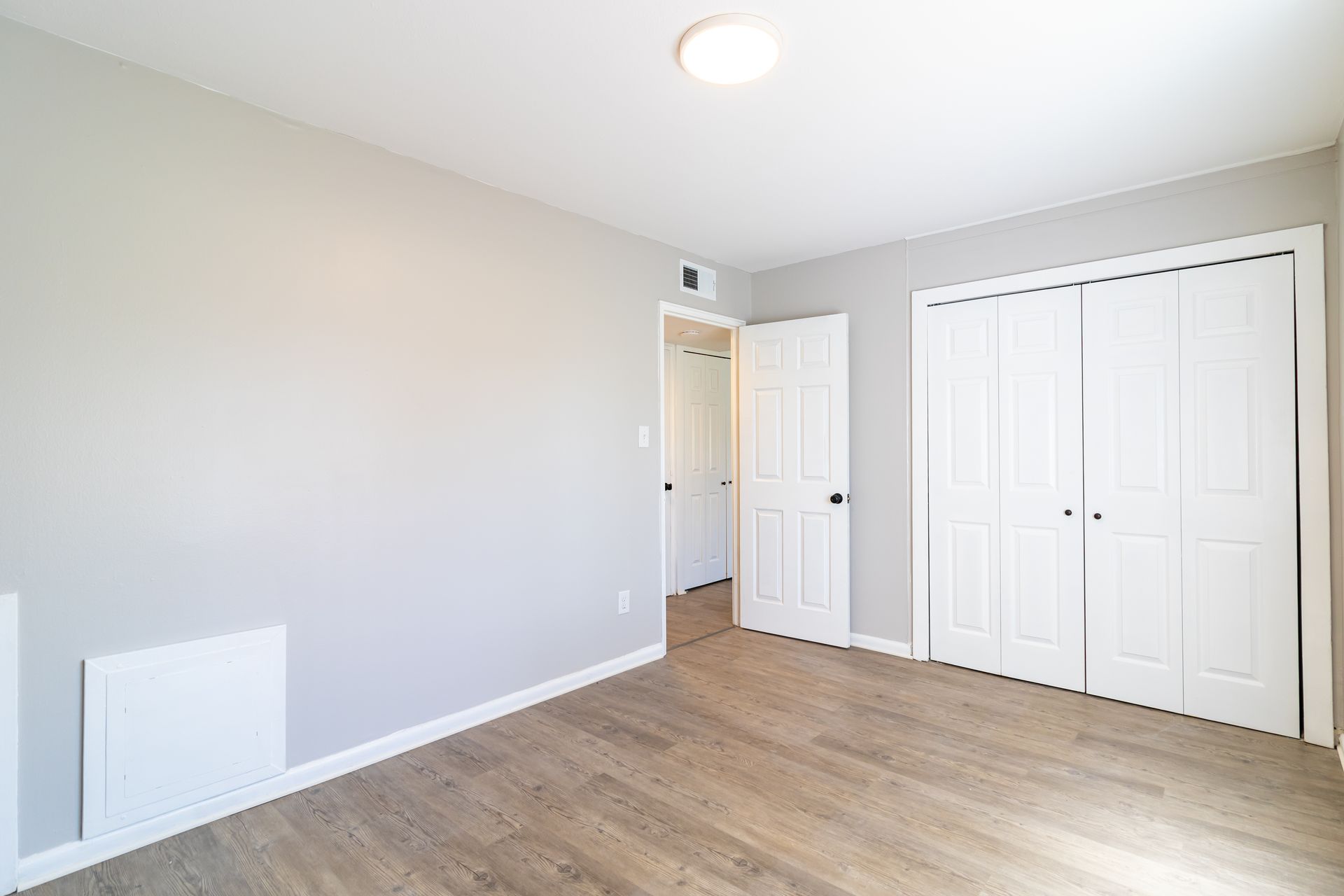 Empty bedroom with light gray walls, white doors, and wood-look flooring.