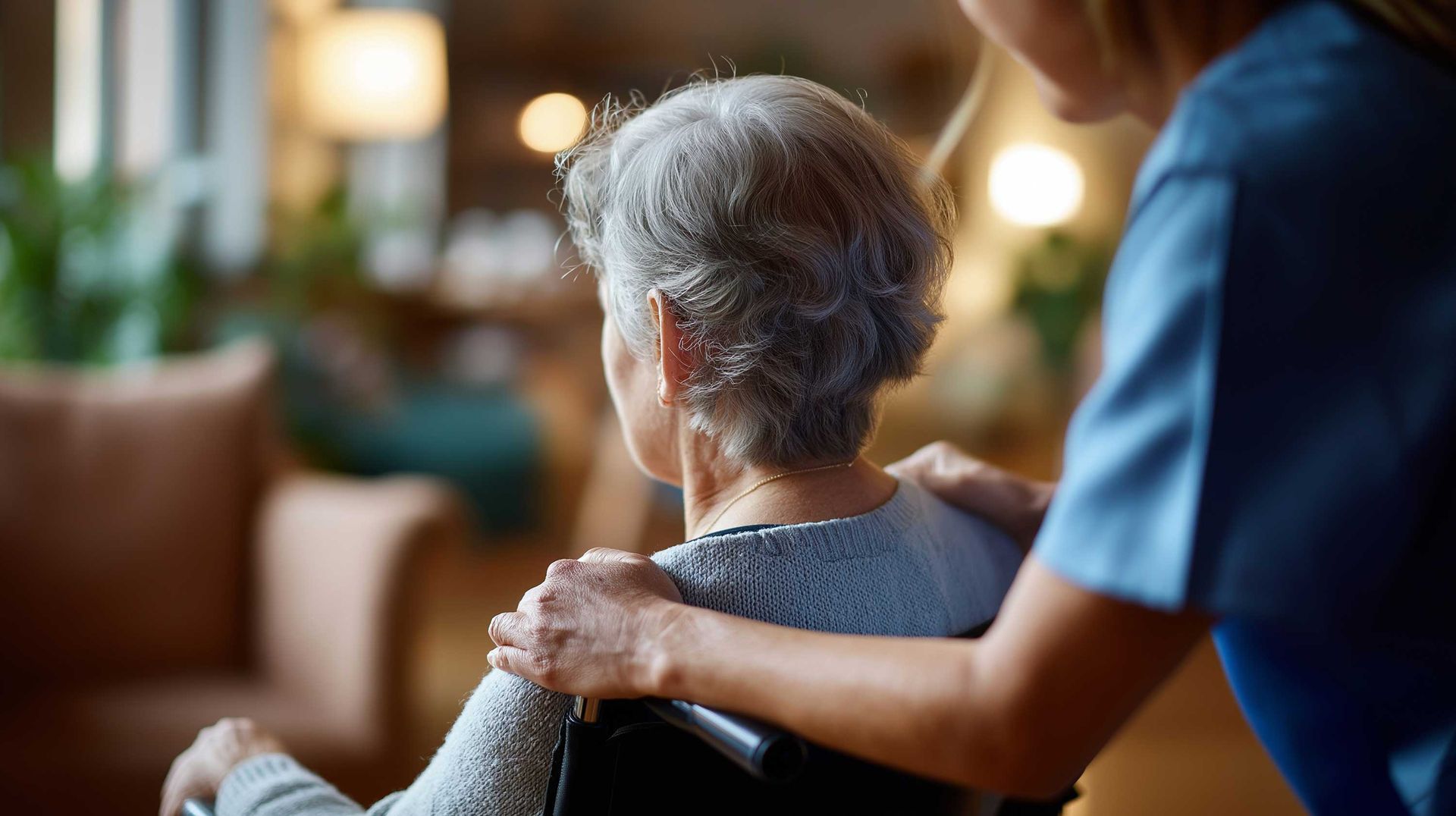 Caregiver standing behind an older adult seated in a wheelchair indoors.