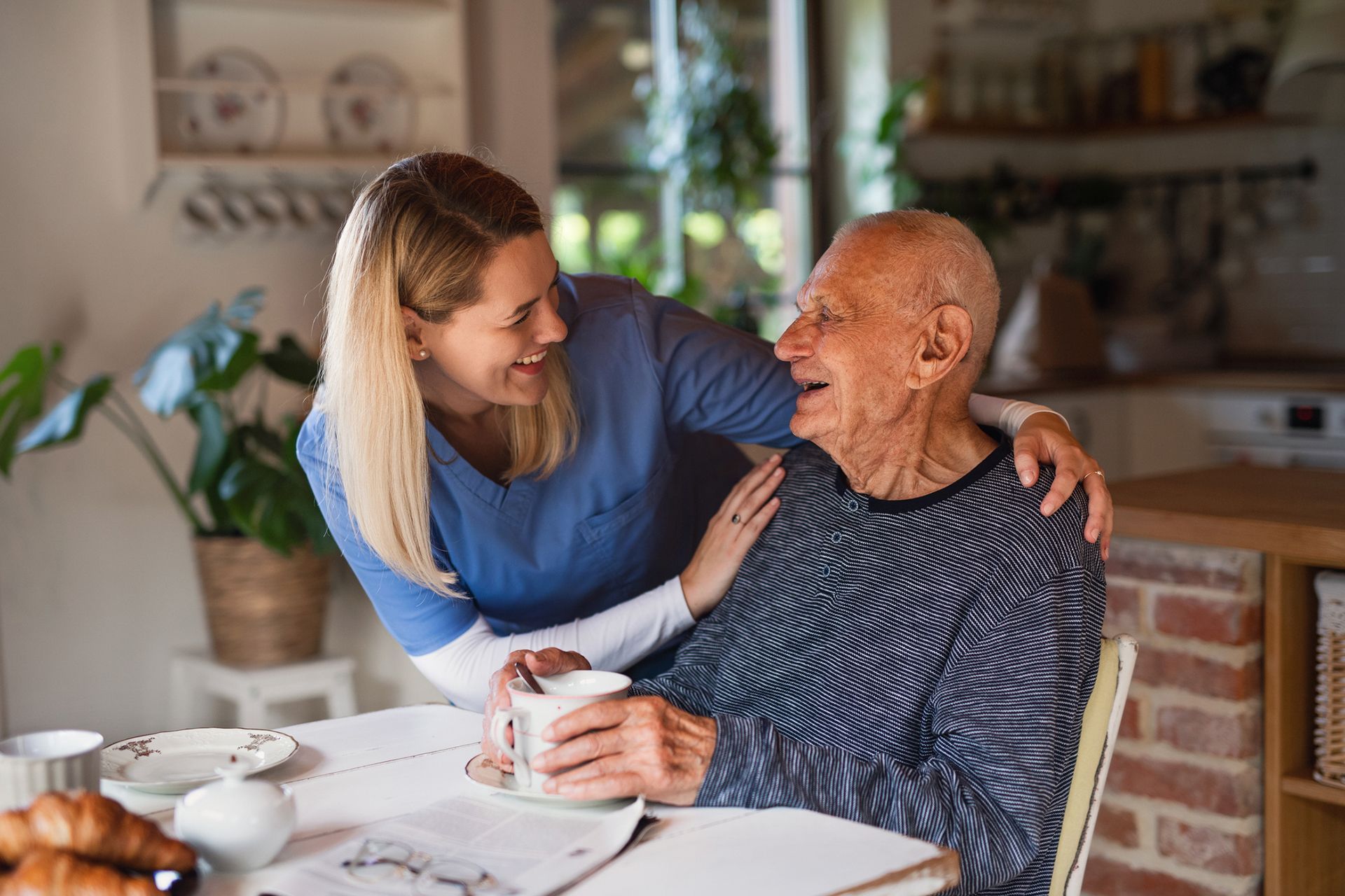 A caregiver smiles, her arm around an elderly man holding a mug at a kitchen table. A caregiver smiles, her arm around an elderly man holding a mug at a kitchen table.