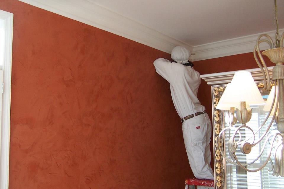 Person in protective suit paints an orange-red wall near white crown molding and a window.
