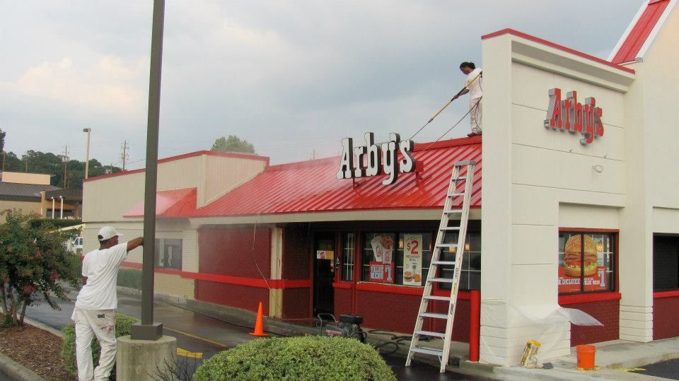 People power washing an Arby's restaurant. One on the roof, one on the ground, both with sprayers, in front of a red and white building.
