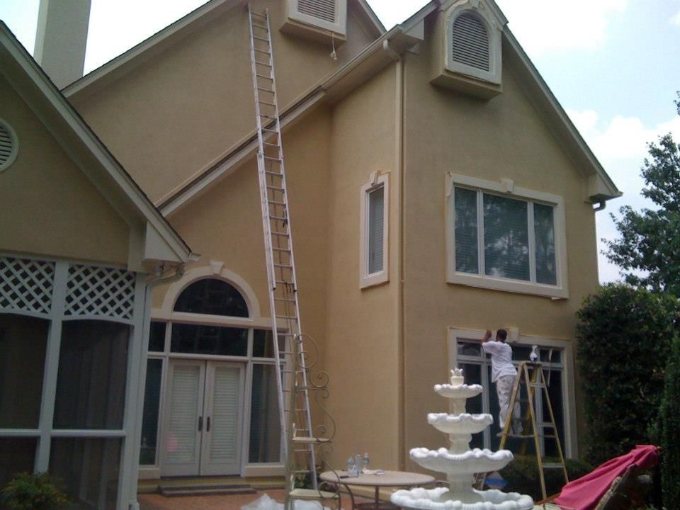 A person painting a tan house exterior, using a tall ladder. A water fountain is in front.