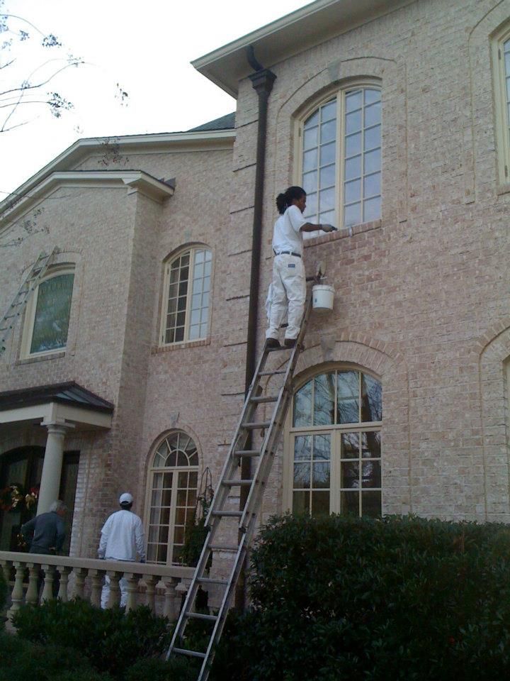 Person painting trim on a two-story brick house, standing on a ladder. Another painter on the balcony below.