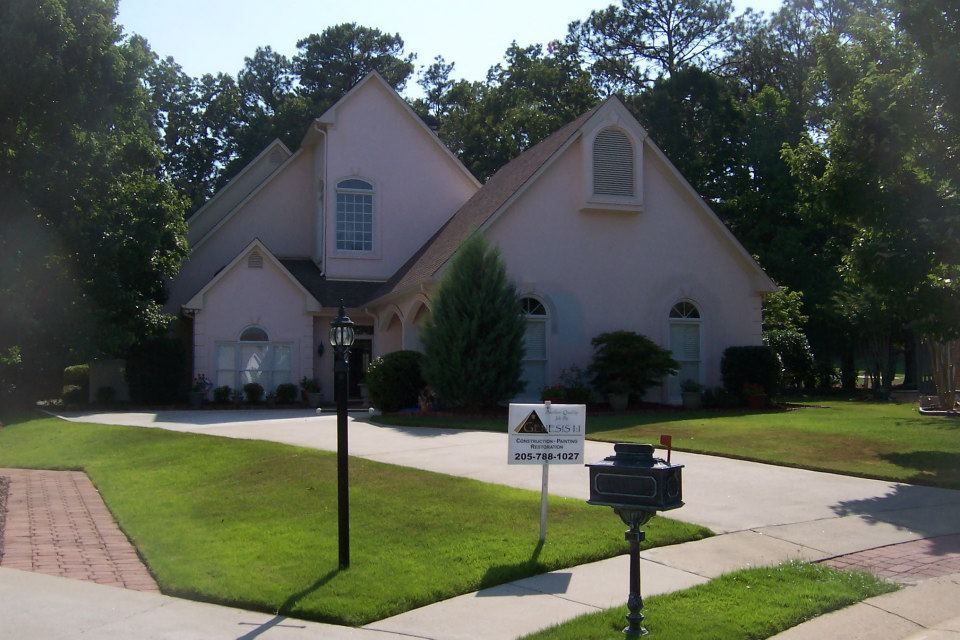 Pink stucco house with a driveway, surrounded by trees and green grass.