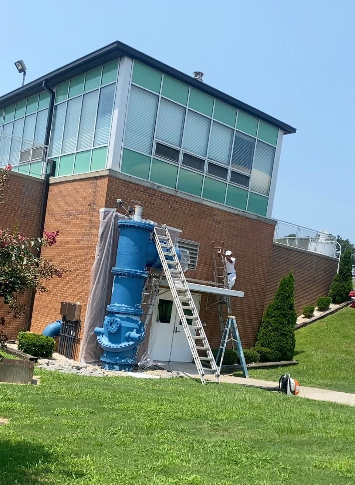 Brick building with blue pipe, a ladder, and a worker in white painting. Green lawn in front.