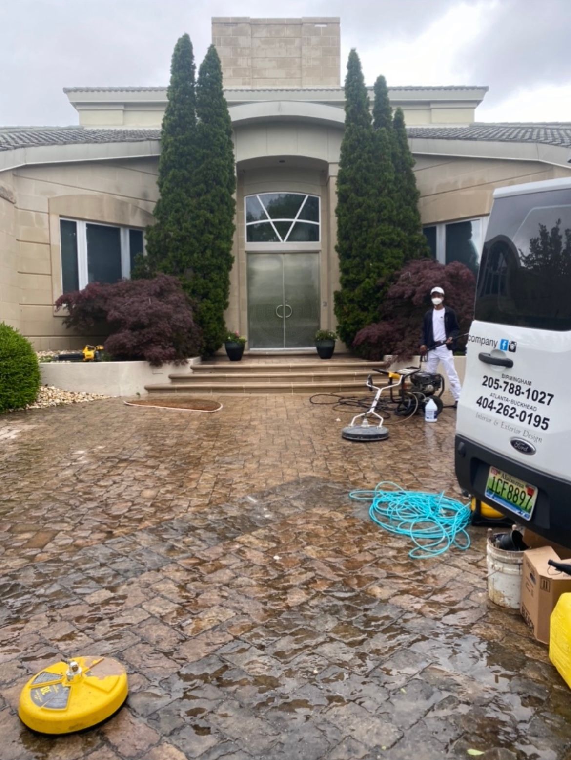 Workers cleaning a paver driveway in front of a large house. A van is parked nearby.