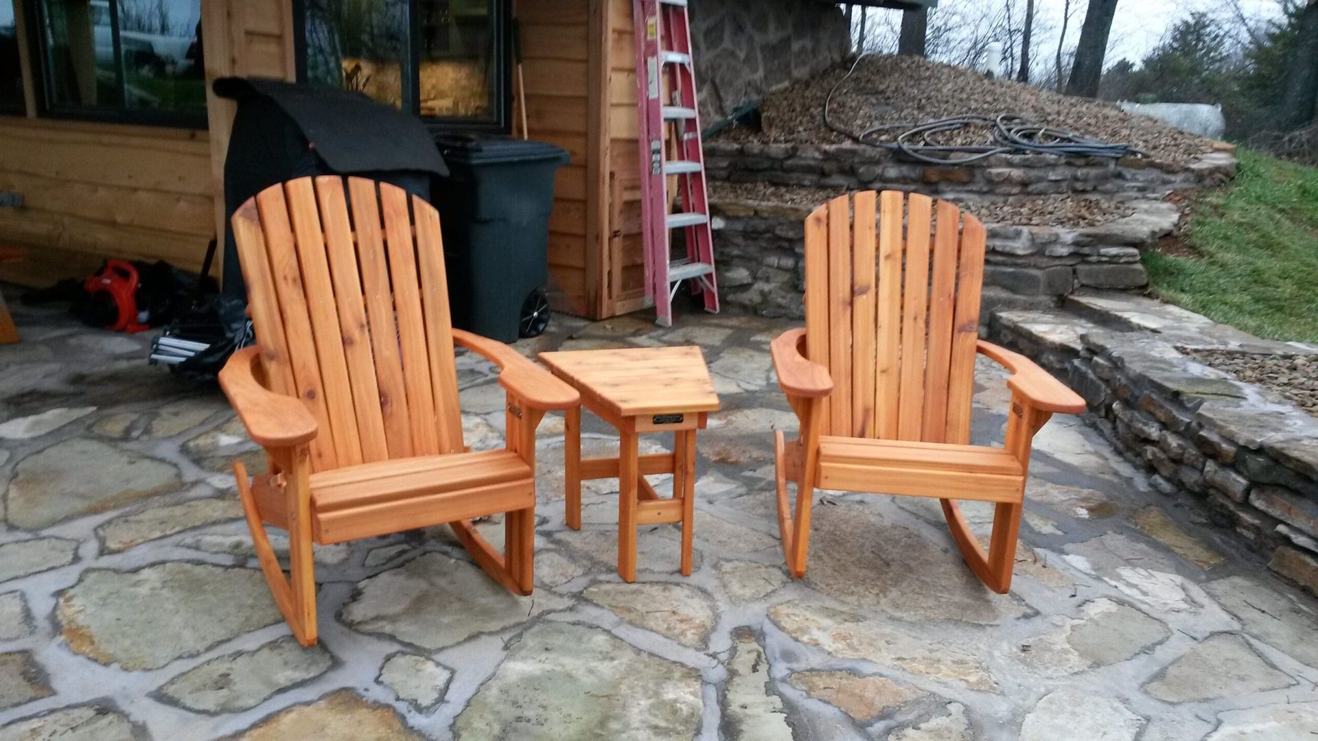 Two wooden adirondack chairs and a table on a stone patio.