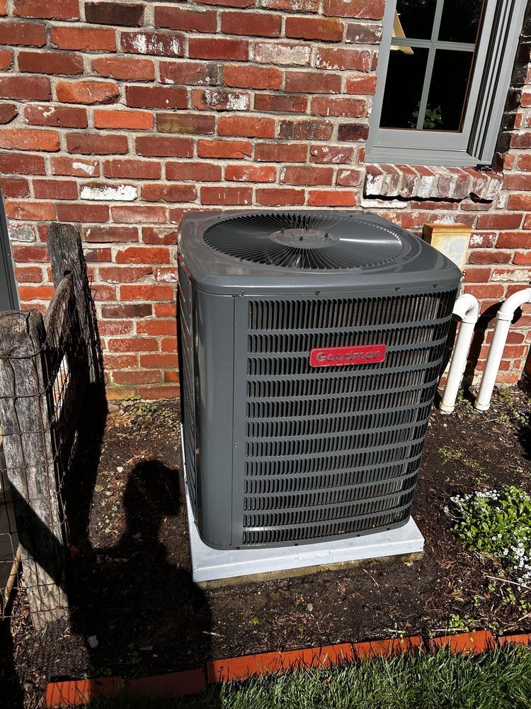 Goodman air conditioner unit against a brick wall. Gray, with a red nameplate.