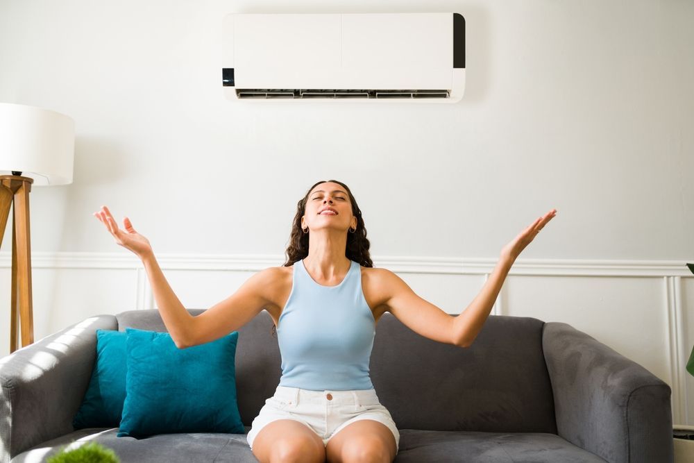 Woman enjoying cool air from AC unit, sitting on couch, arms raised.