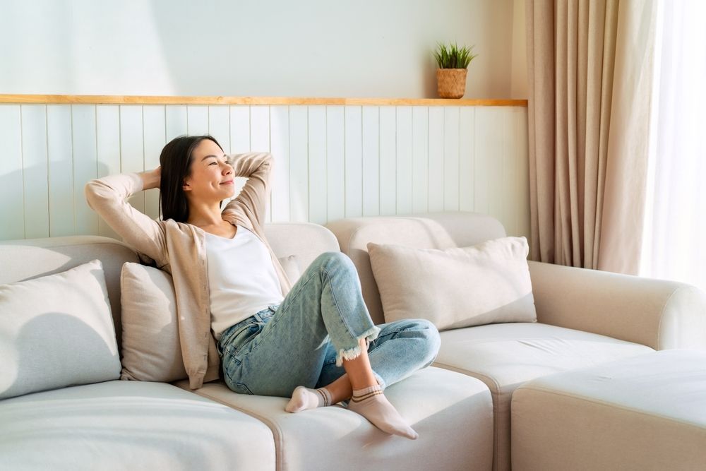 Woman relaxes on a light-colored couch, smiling with her hands behind her head, in a sunlit living room.