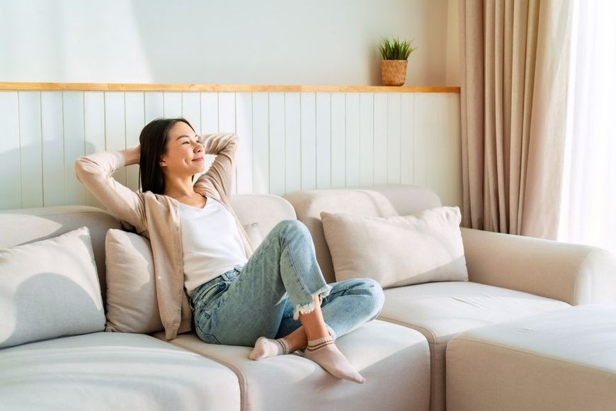 Woman relaxes on a light-colored couch, smiling with her hands behind her head, in a sunlit living room.