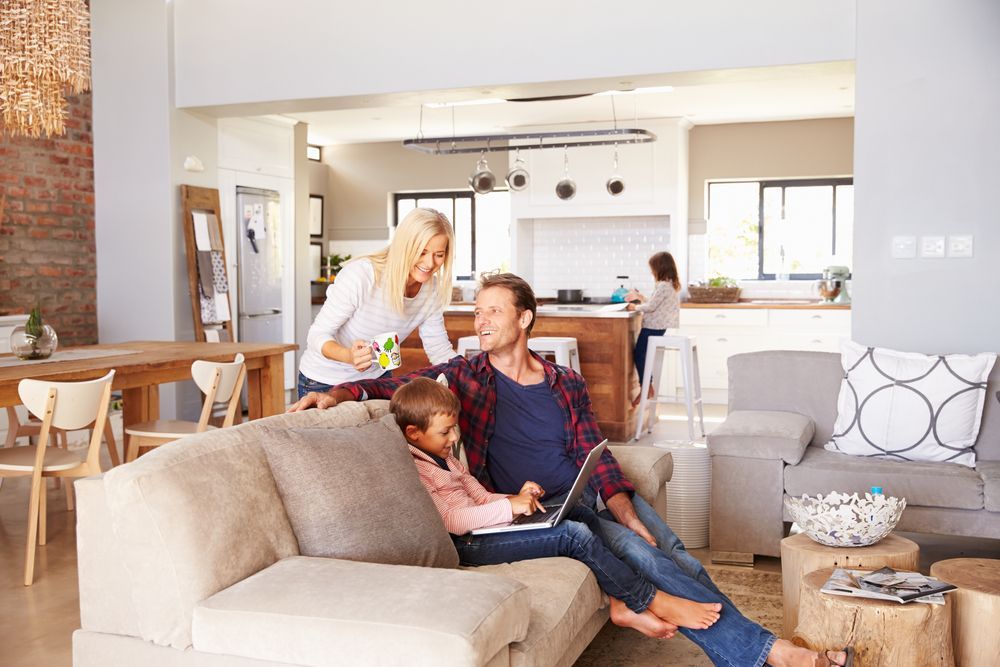 Family in living room: Child and parent on sofa with laptop, another parent holding mug, person in kitchen.