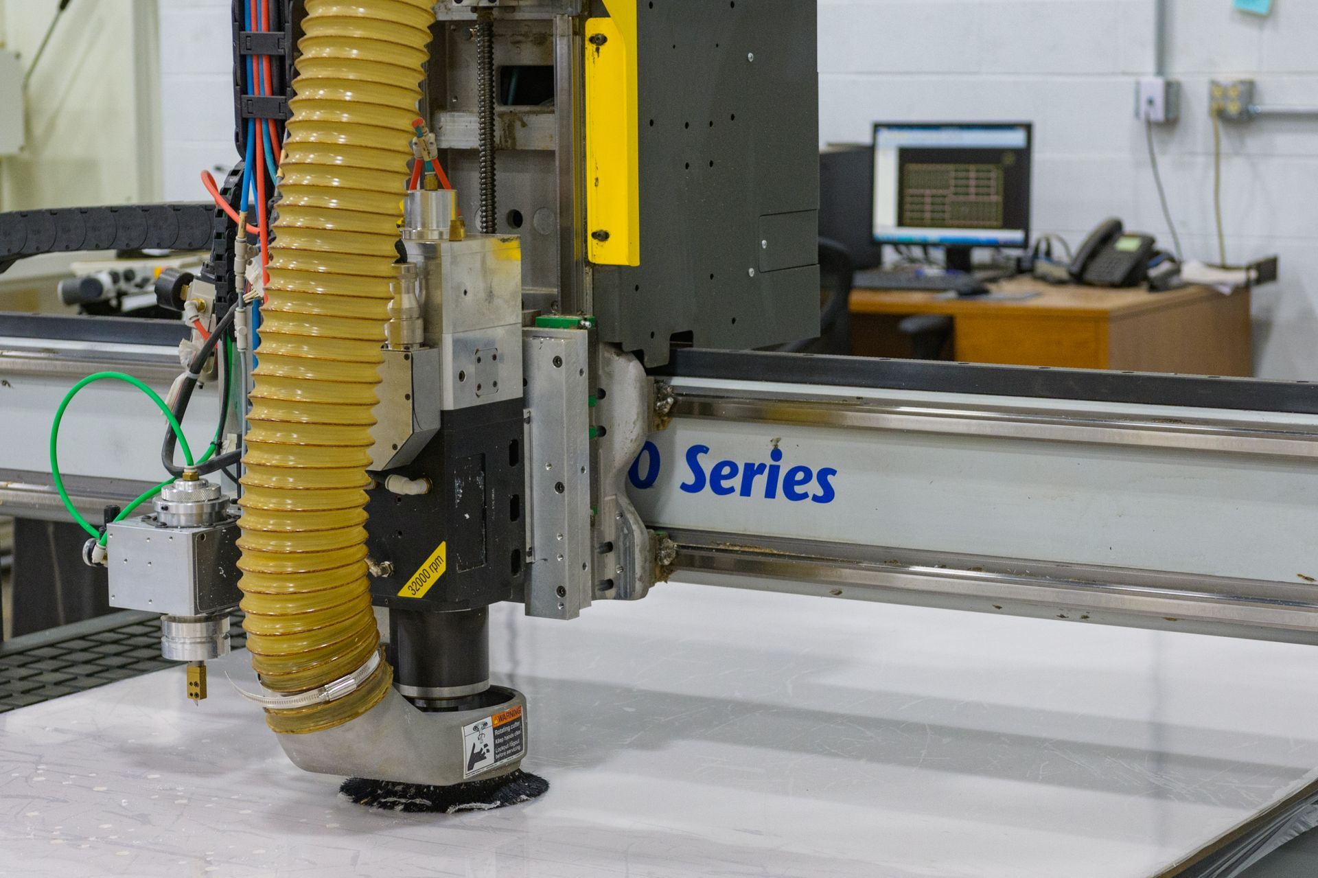 A CNC router machine cutting into a large, white flat material in a workshop, with a yellow vacuum hose attached.