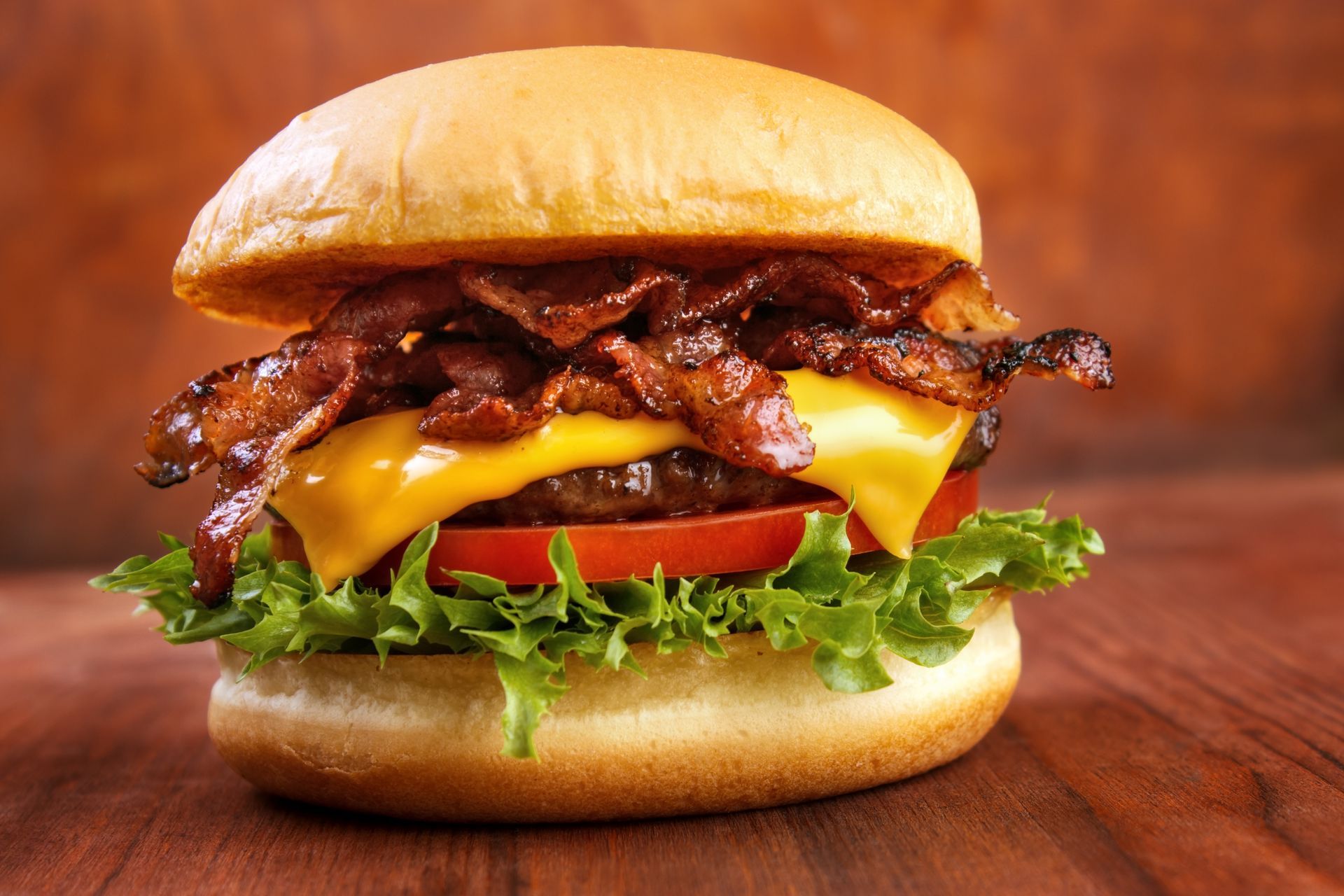 A hamburger with cheese , bacon , lettuce and tomato on a wooden table.