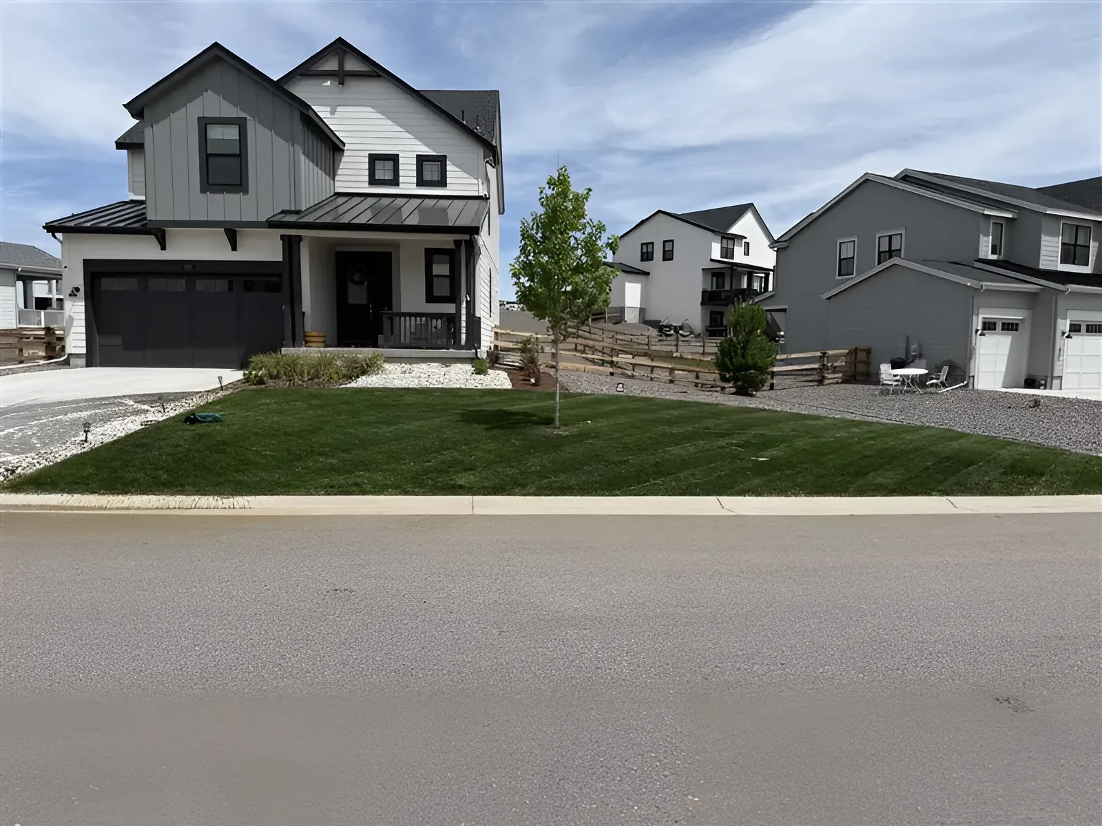 Two-story house with green lawn, young tree, and other homes in the background under a blue sky.