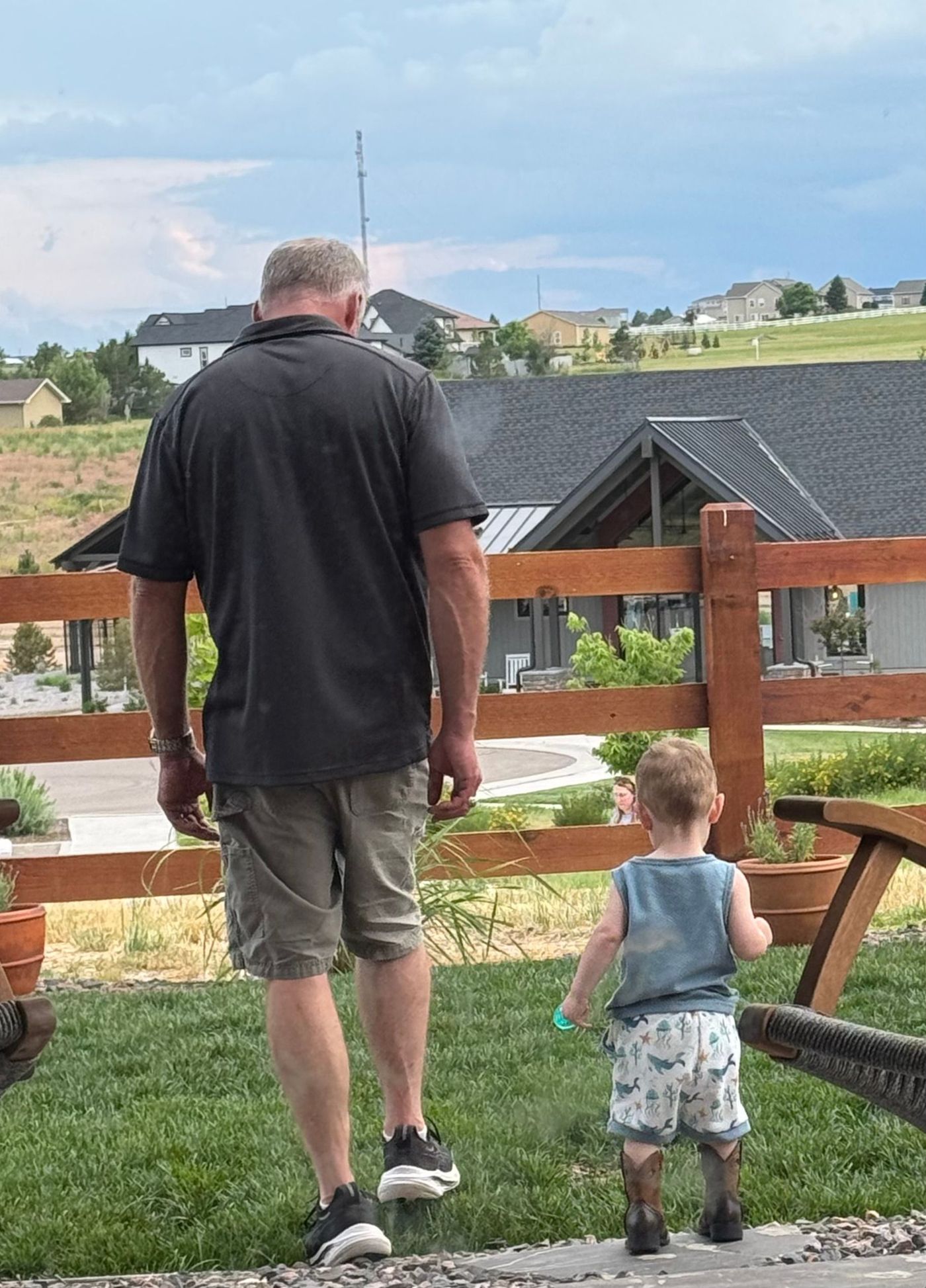 A man and a toddler walk on grass near a wooden fence, houses in the background.