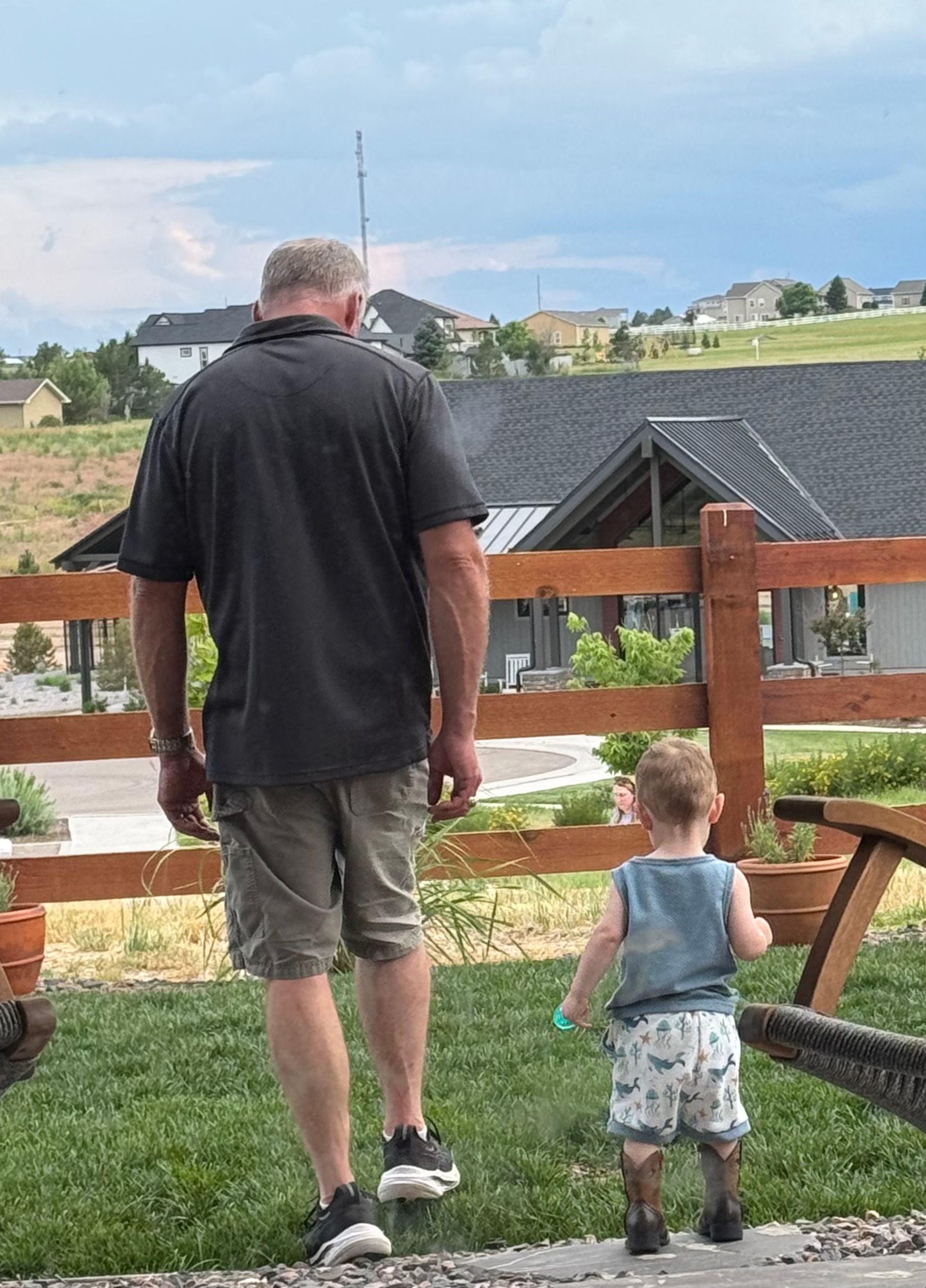 A man and a toddler walk on grass near a wooden fence, houses in the background.