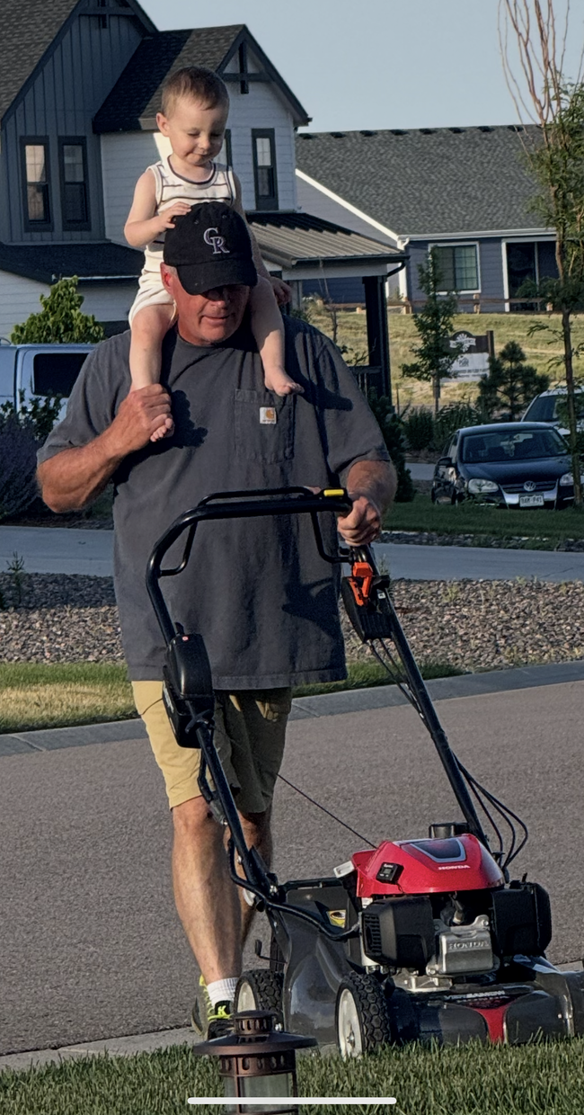 Man mowing lawn with child on his shoulders. Suburban setting, sunny day.