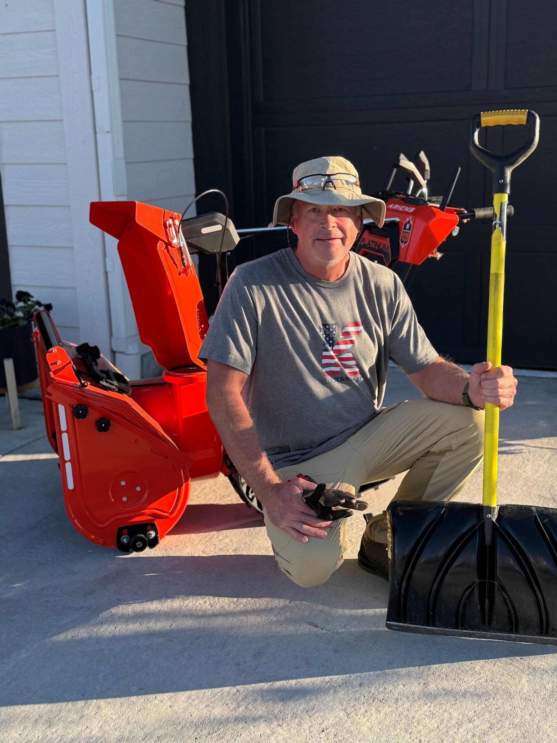 Man kneels beside snowblower and shovel. He wears a hat and t-shirt with an American flag. Outdoors.