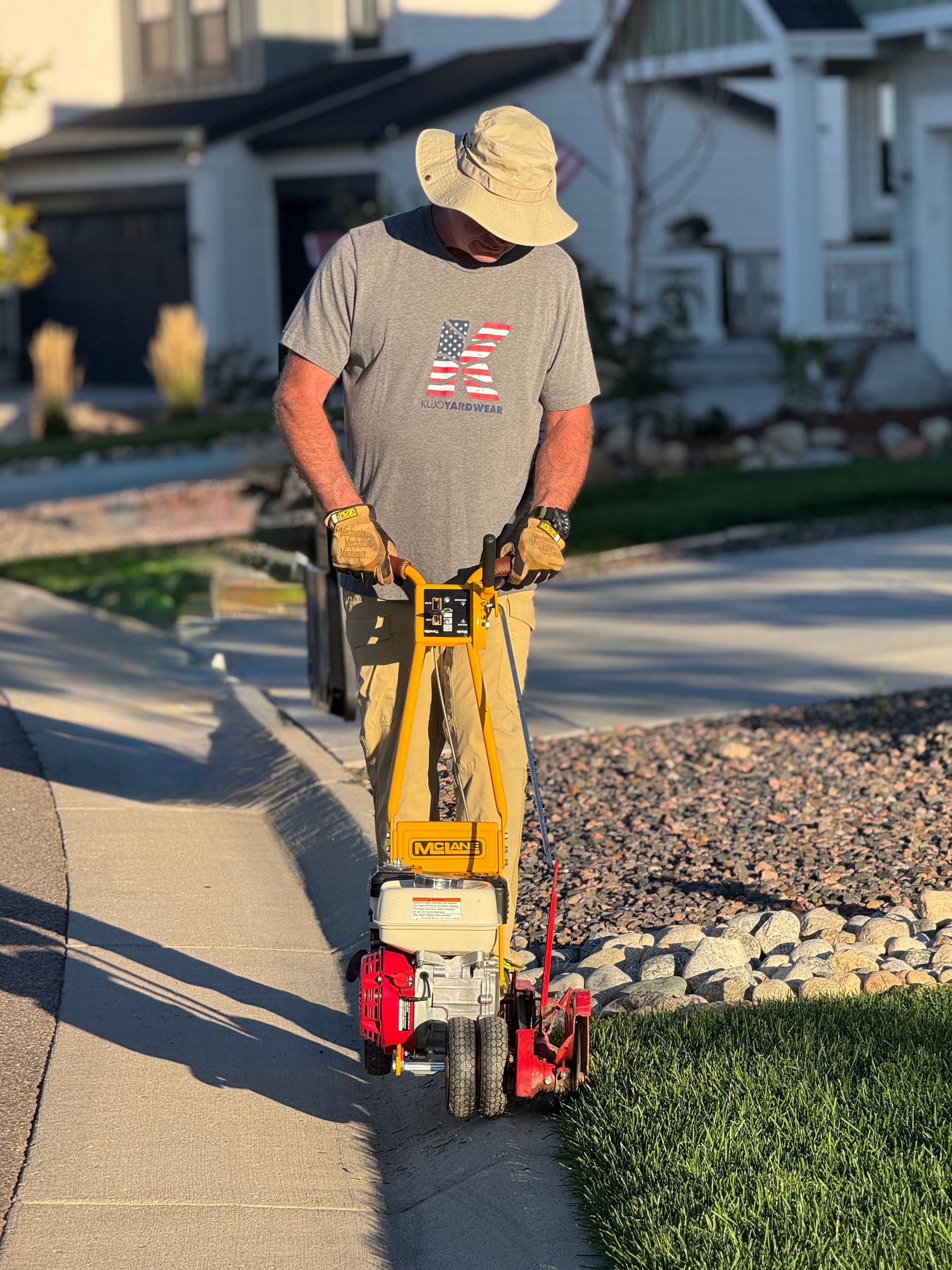 Man using a lawn edger to trim grass alongside a sidewalk. He wears a hat and gloves.