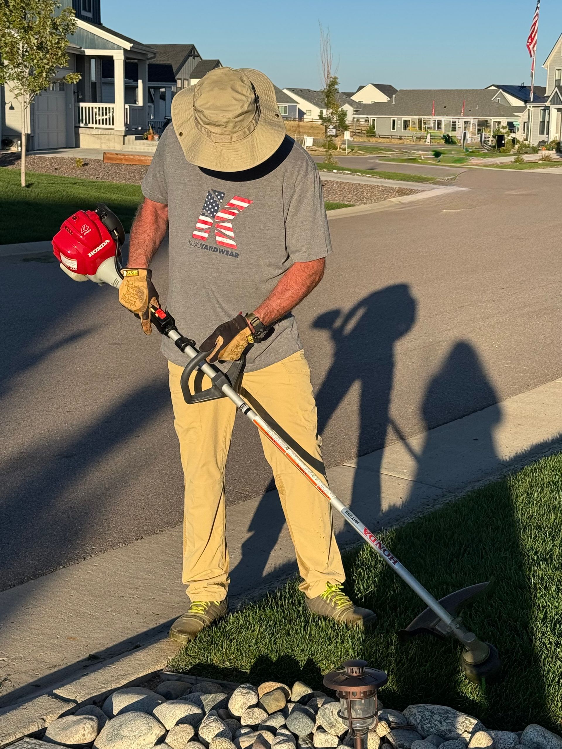 Man using a string trimmer on grass border near a street, wearing a hat, tan pants, and a gray t-shirt.