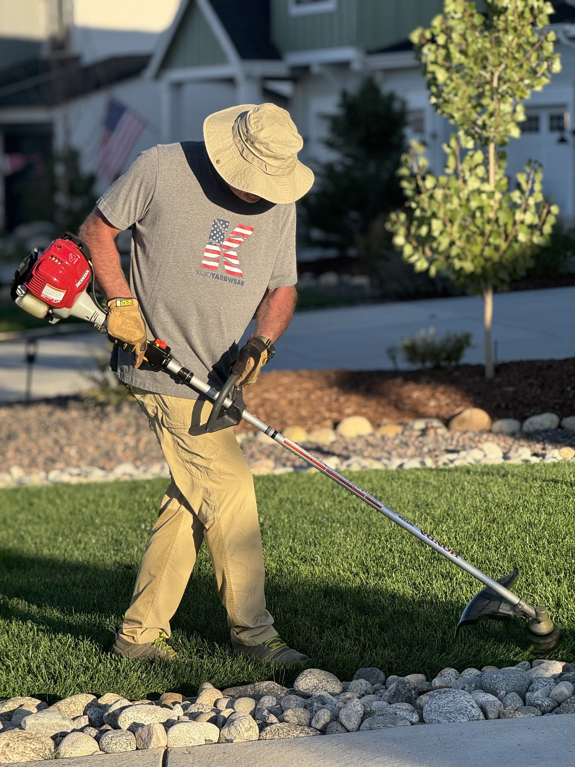 Man in hat trimming grass with a weed wacker in front of a house.