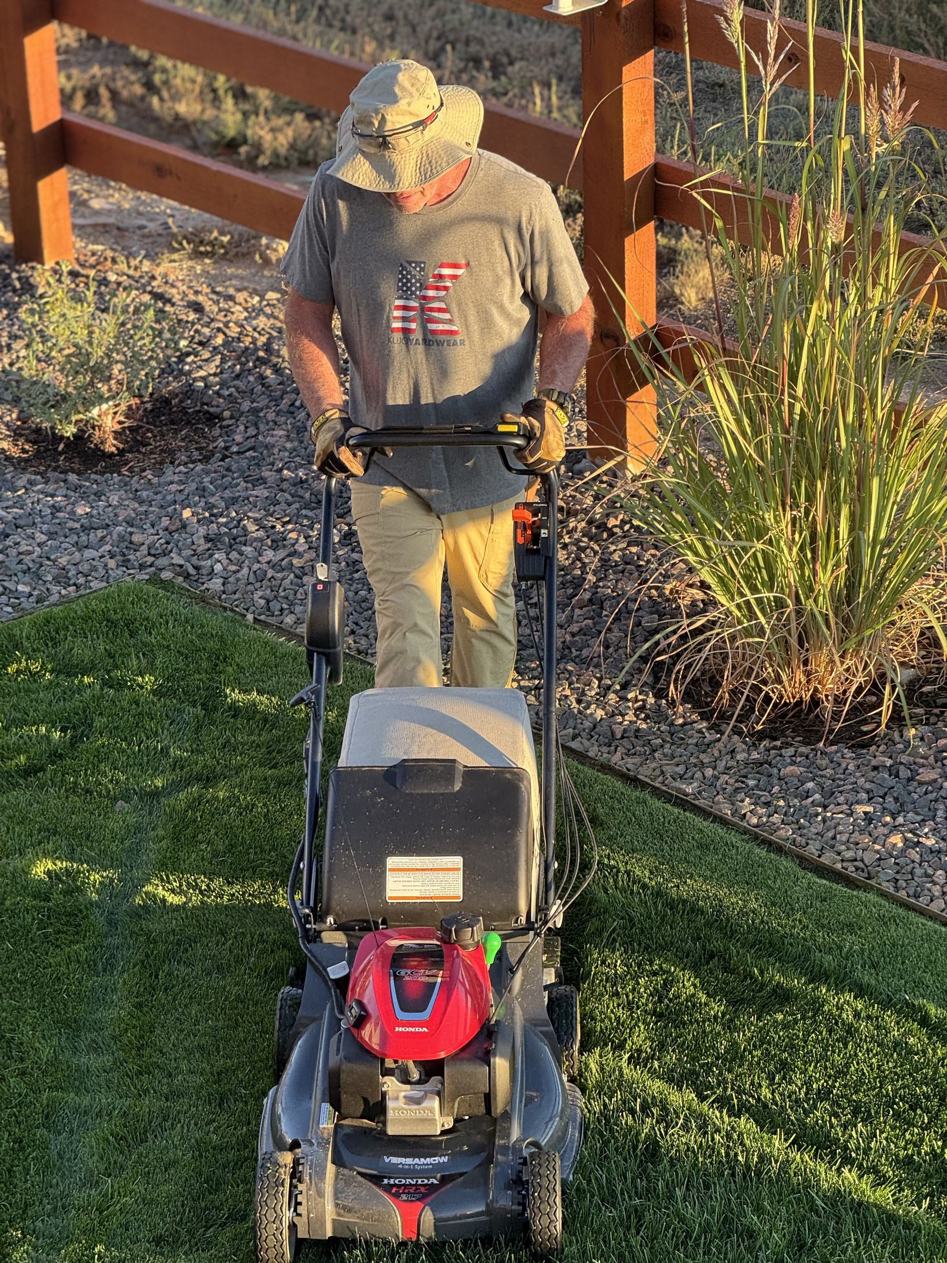 Man mowing a green lawn with a red and black lawnmower in a yard, wearing a hat and gloves.