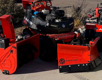 Man spraying lawn with a backpack sprayer in front of a house, late afternoon light.