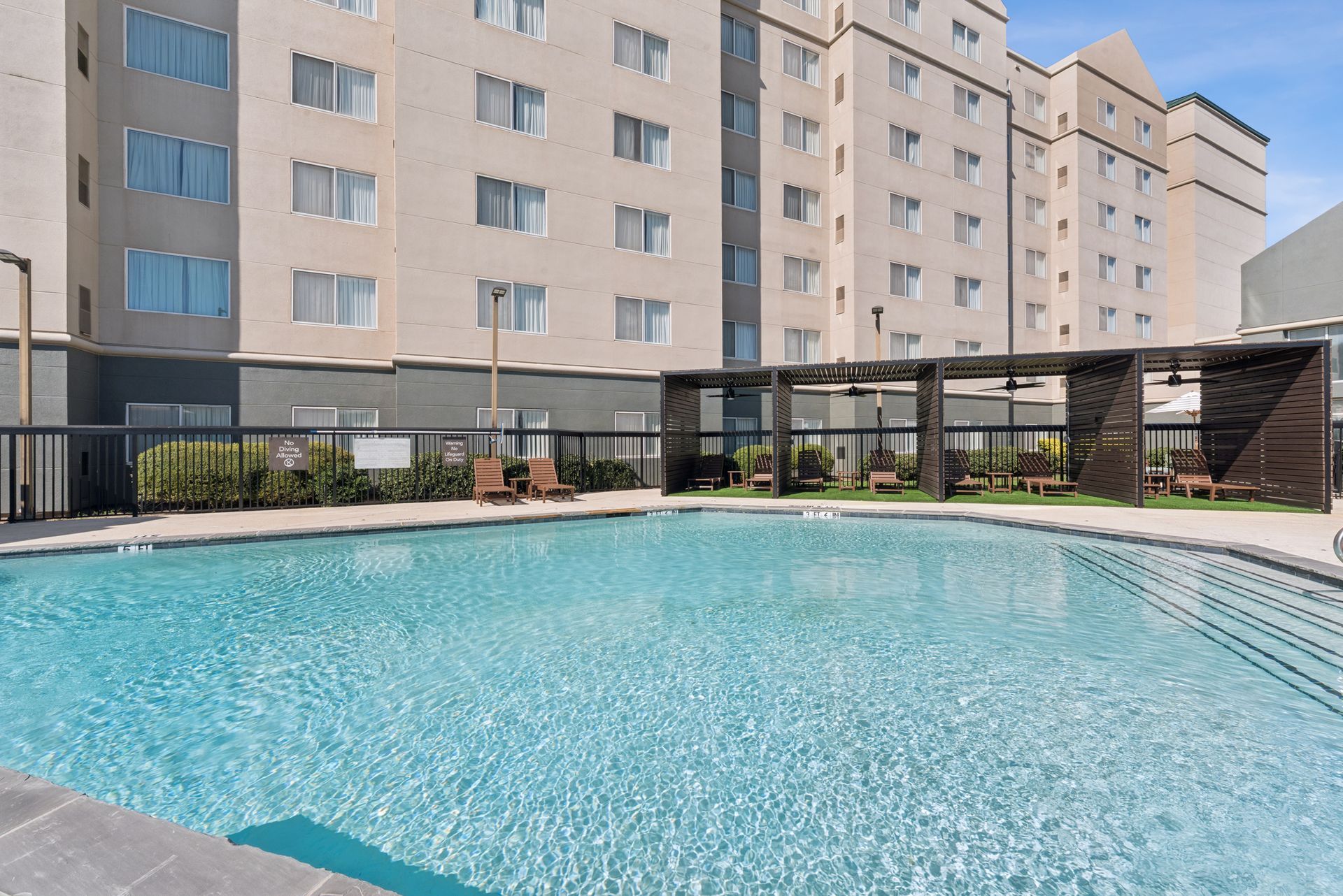 Swimming pool in front of a tall building with lounge chairs and a covered seating area.