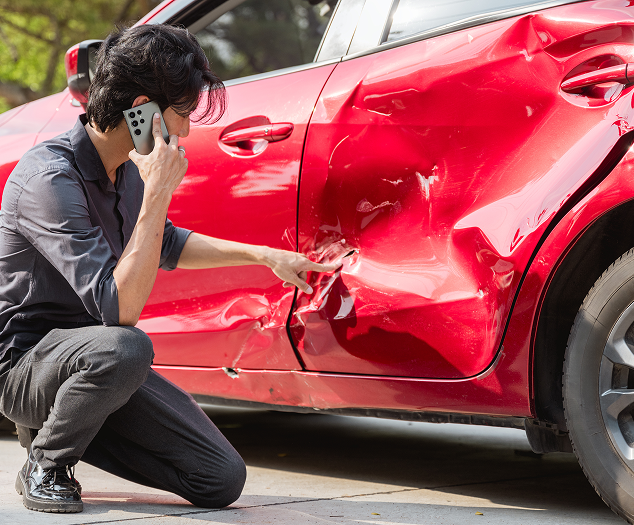 Man on phone kneels by red car with significant damage to the side | CARSTAR Oakville West