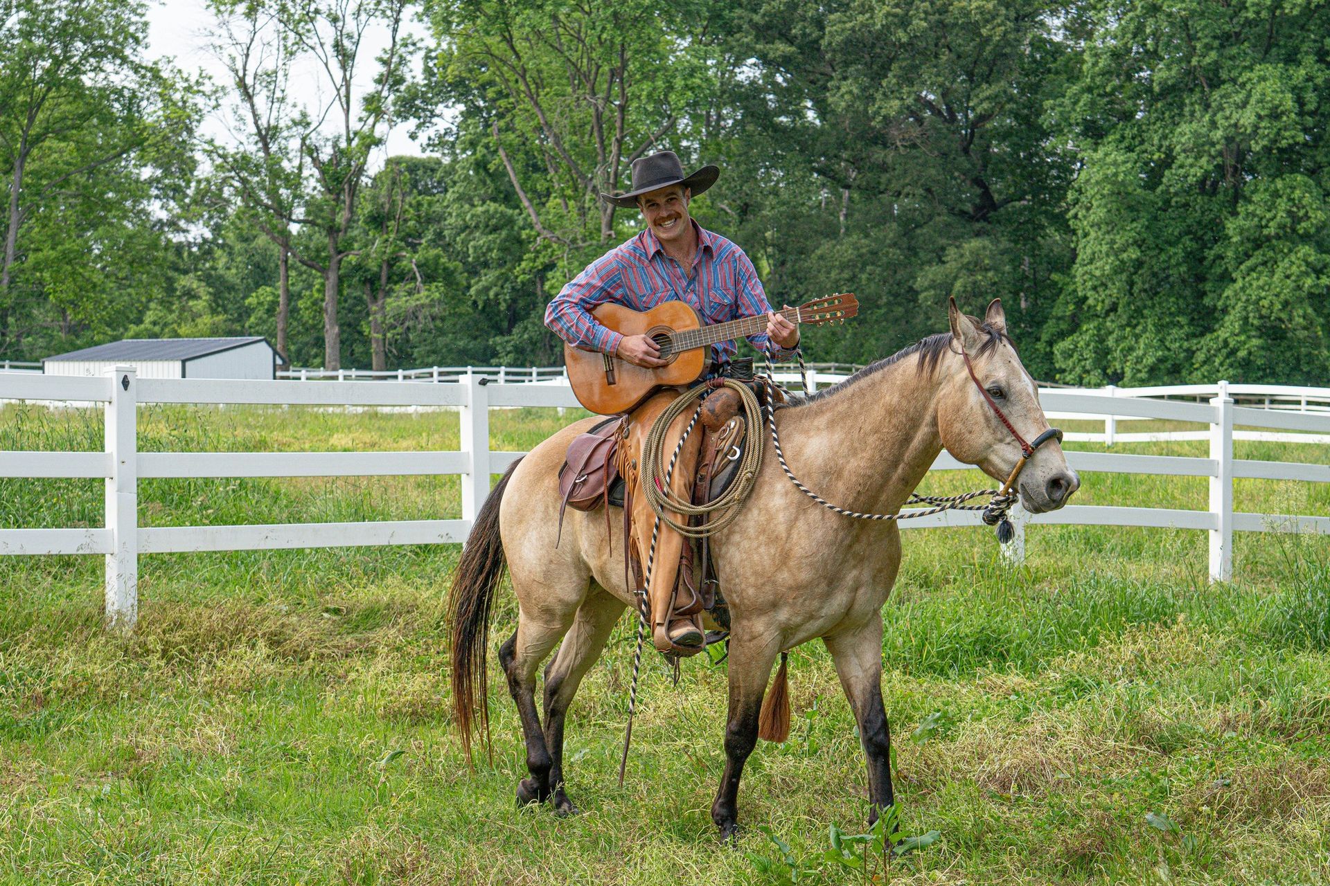 Man in cowboy hat playing guitar while riding a horse in a pasture.