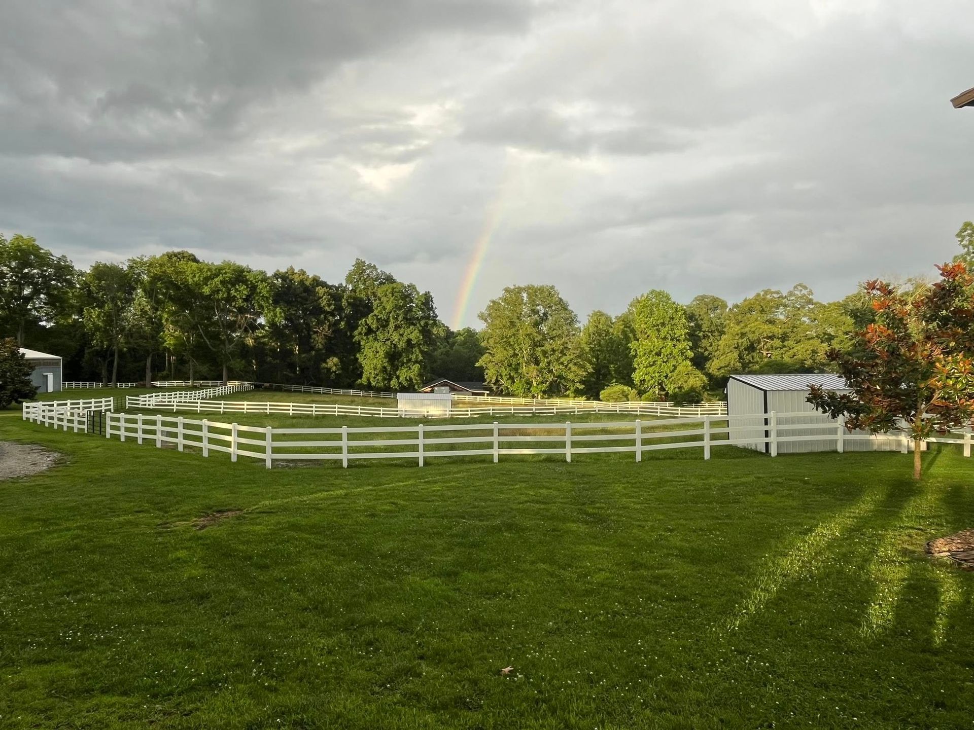 A rainbow arcs over a white-fenced pasture, with trees, pond, and a shed under a cloudy sky.