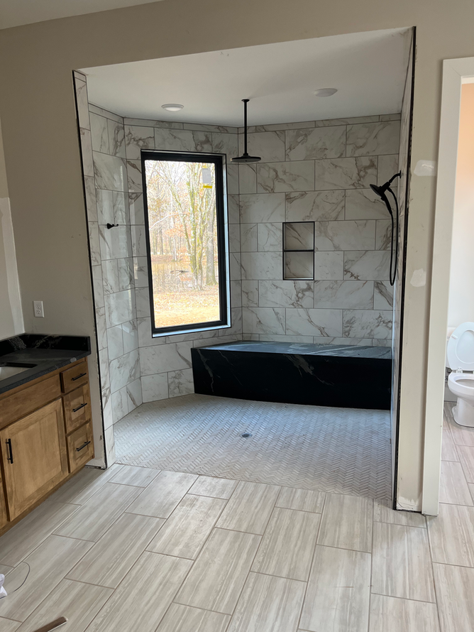 Bathroom with marble-tiled shower, black tub, and large window overlooking trees.