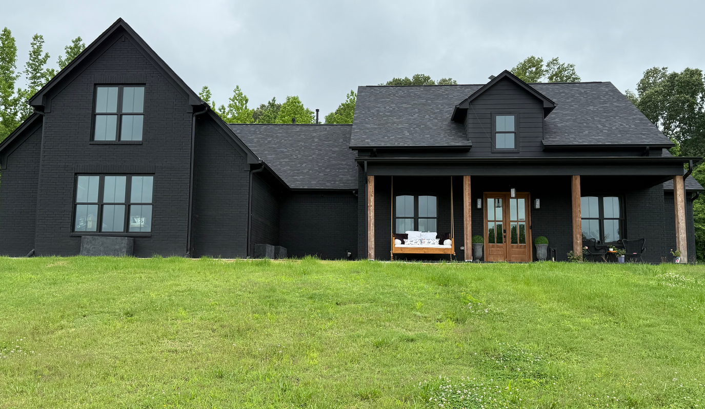 A black house with a porch is sitting on top of a lush green field.