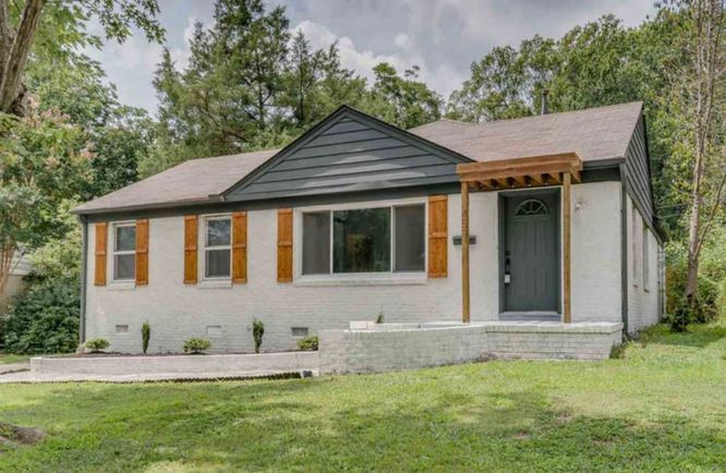 A white house with a black roof and orange shutters is surrounded by trees.