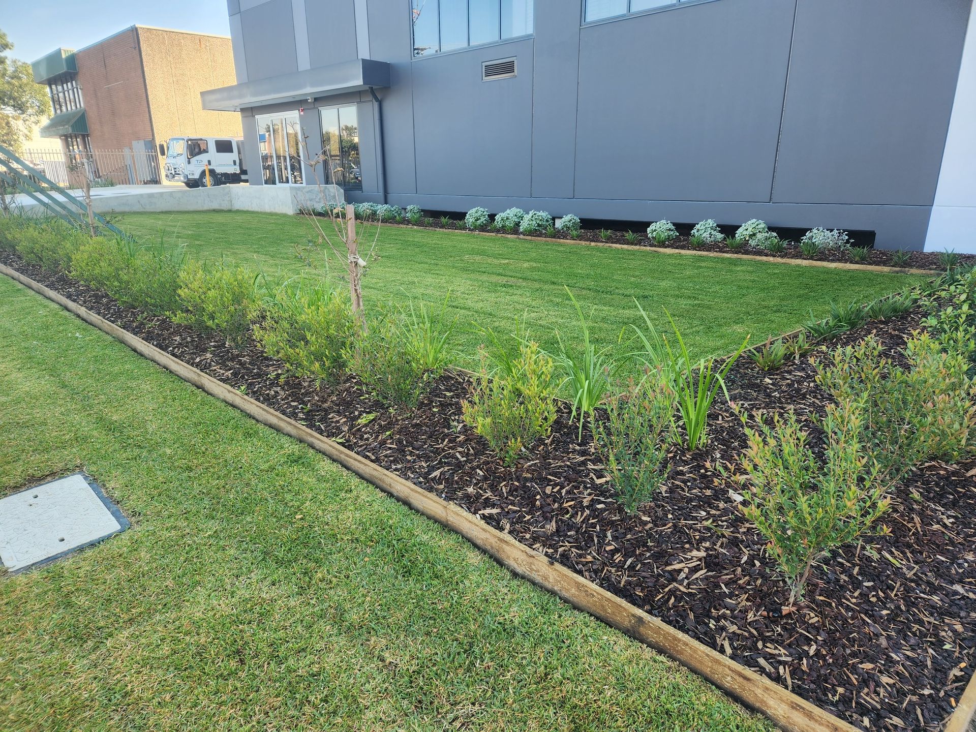 A Landscaped Lawn with Shrubs and Mulch Borders a Gray Building — Total Perfection In Kariong, NSW