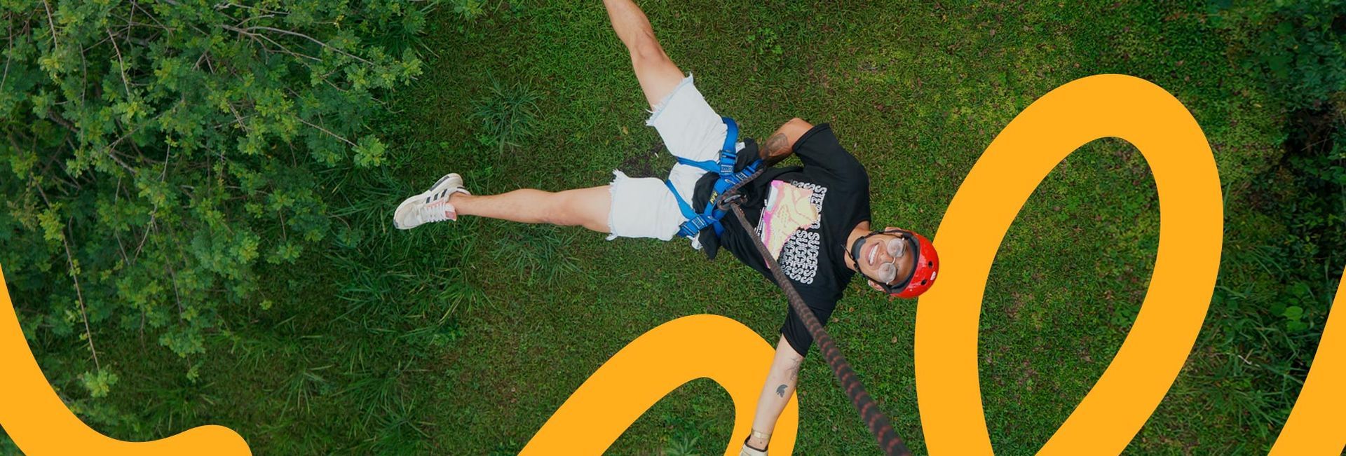 Persona haciendo puenting sobre un paisaje verde, vistiendo un casco rojo y pantalones cortos blancos.