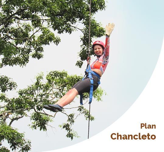 Mujer descendiendo por una cuerda, saludando, con casco y arnés en un entorno forestal.