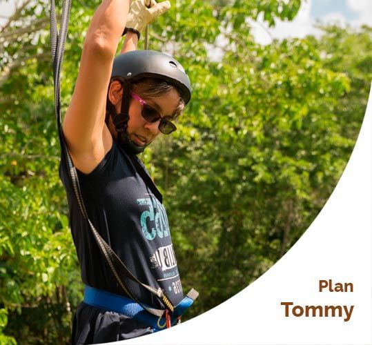 Persona en una tirolina con casco, gafas de sol y arnés, rodeada de árboles.