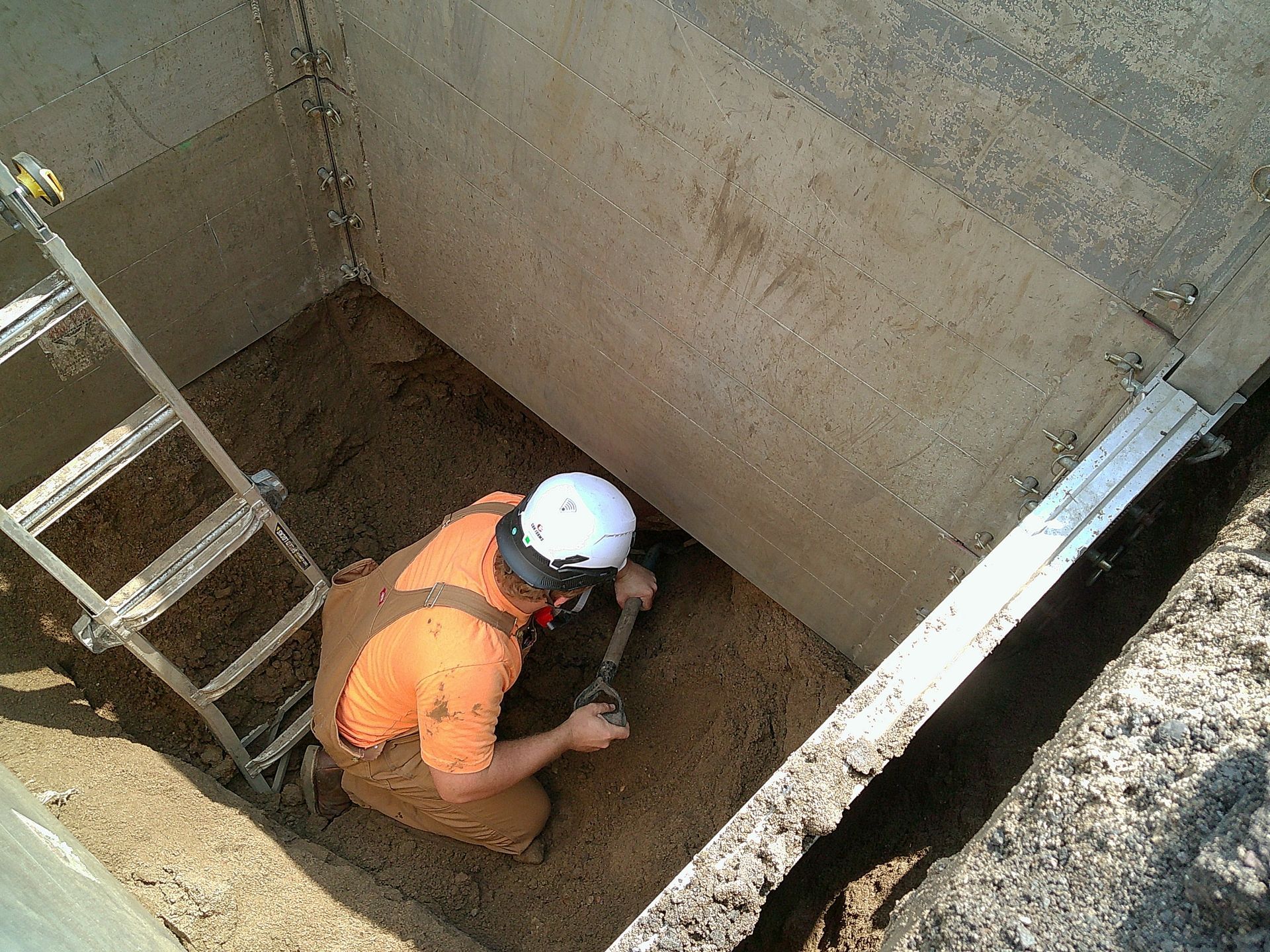 Person with hard hat excavating inside concrete structure, using a shovel and near a ladder.